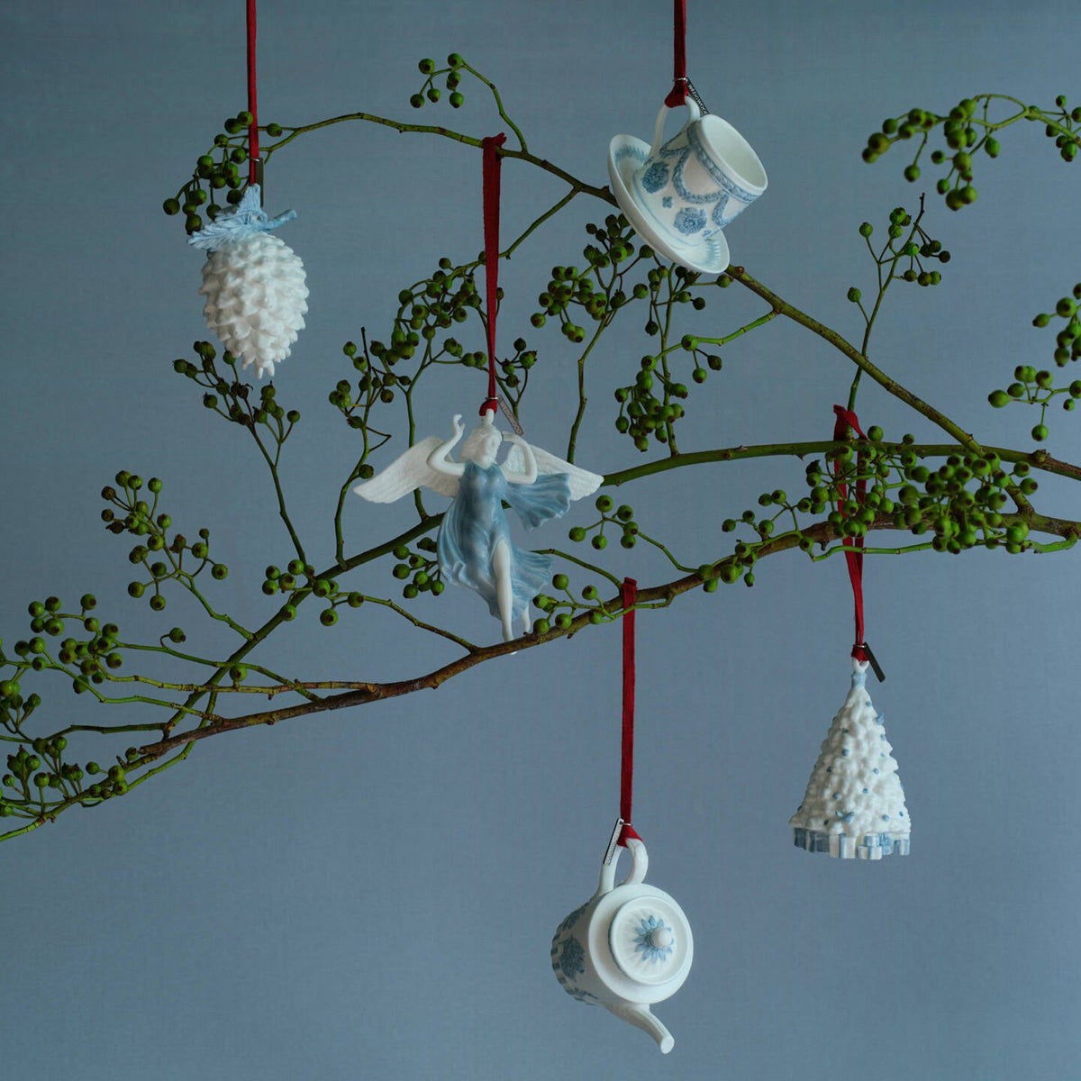 Five ceramic Wedgwood blue and white Christmas ornaments shaped like a pinecone, angel, teapot, teacup, and miniature Christmas tree, hanging from a decorative branch with red strings against a blue-gray background.