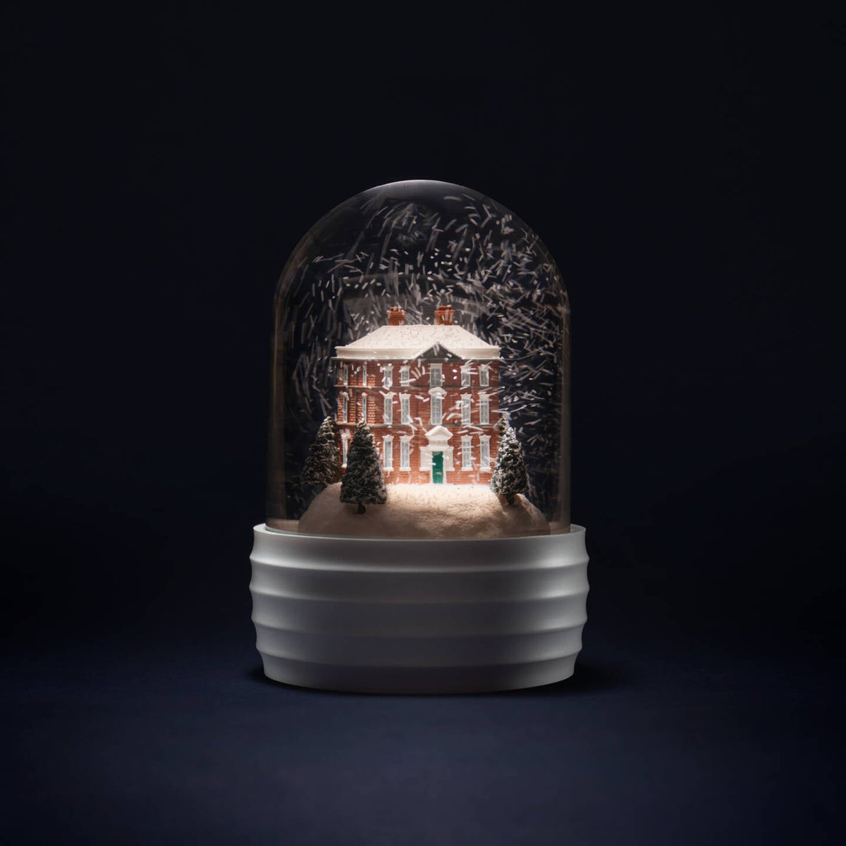 A decorative Wedgwood house glass dome snow globe on a ribbed white base, featuring a miniature red brick house surrounded by snow and trees, photographed against a very dark studio background.