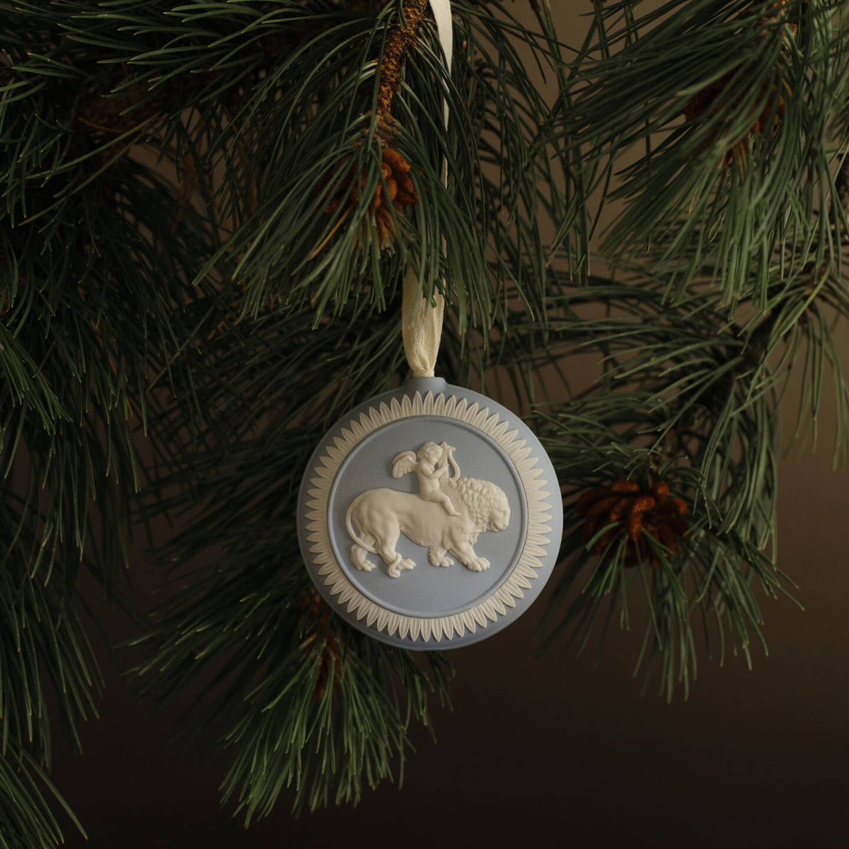 A round decorative hanging Wedgwood jasperware ornament with a lion and lamb motif, suspended from a green pine tree branch against a dark brown background.