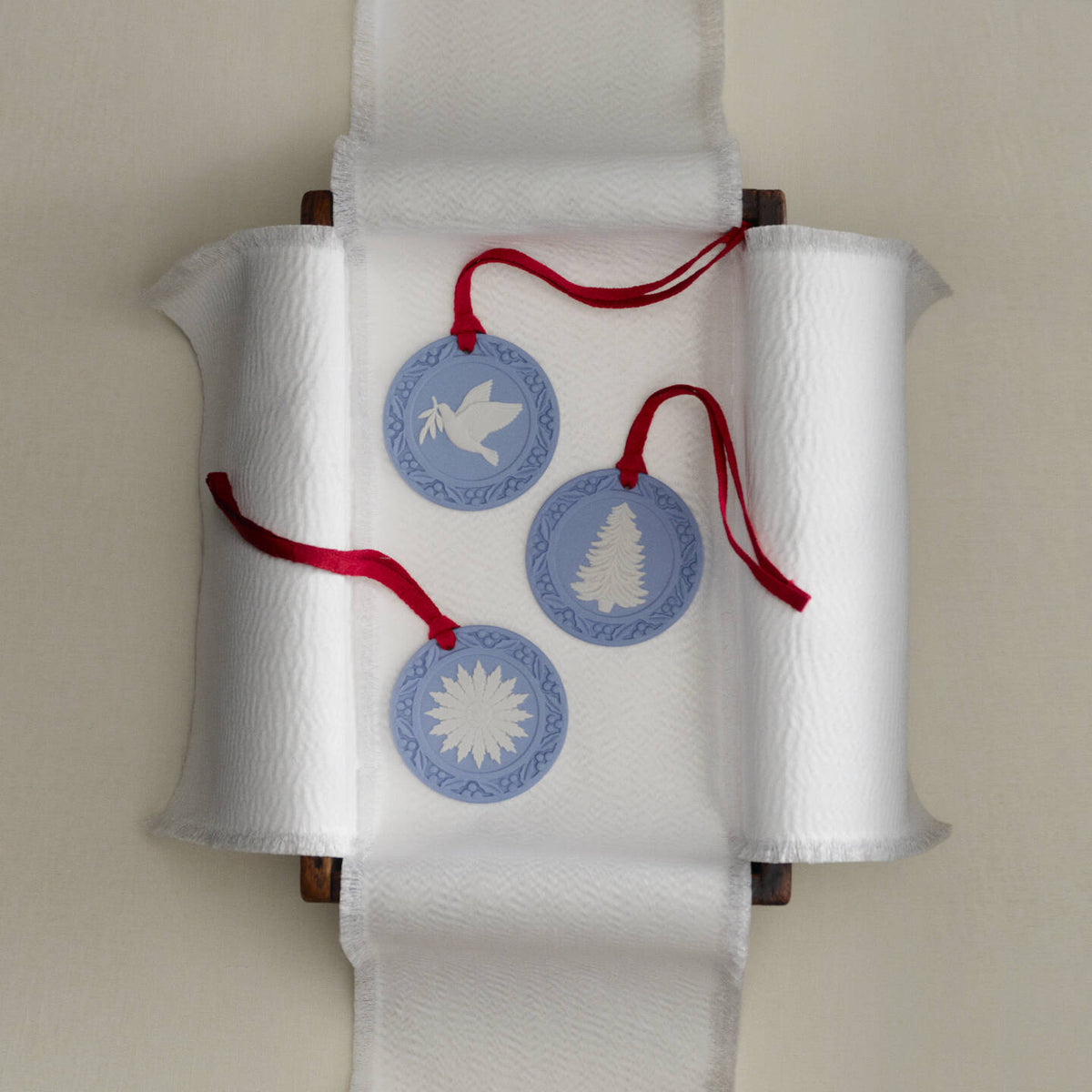Three round Wedgwood jasperware blue and white decorative hanging ornaments with red hanging strings, displayed on a white paper towel roll holder against a light beige wall.