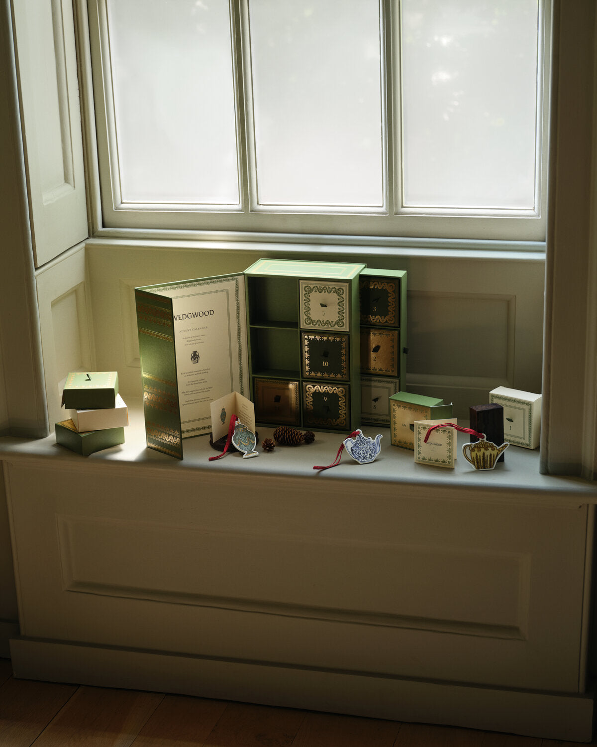 A window sill displays a curated collection of decorative items, including a large green and gold Wedgwood icons advent calendar, including miniature teapots and vase hanging decorations. 
