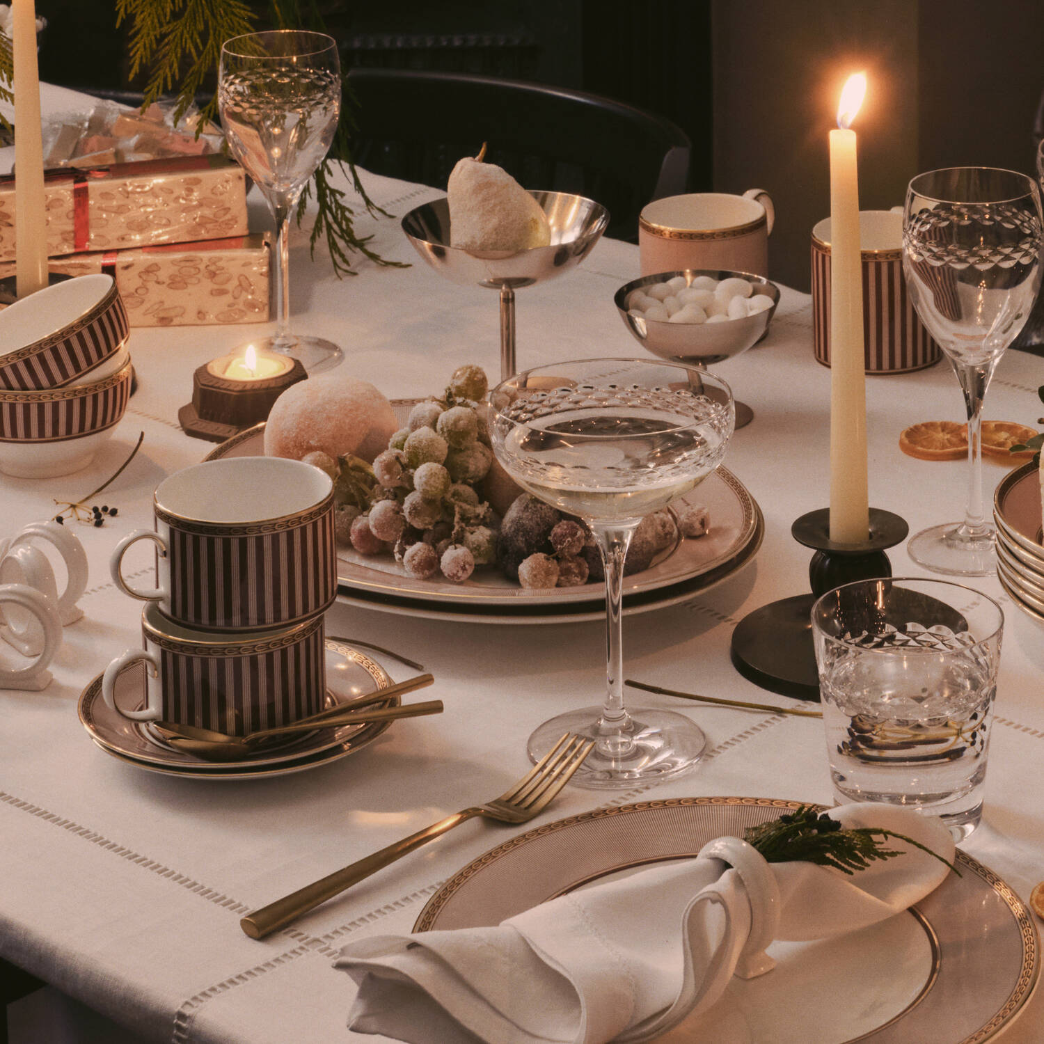 A warmly lit table setting with stacked Wedgwood Red Splendour ceramic mugs in burgundy and cream stripes, crystal-style drinking glasses, candlesticks, plates, napkins, and a centerpiece of grapes and fruit on a white tablecloth.