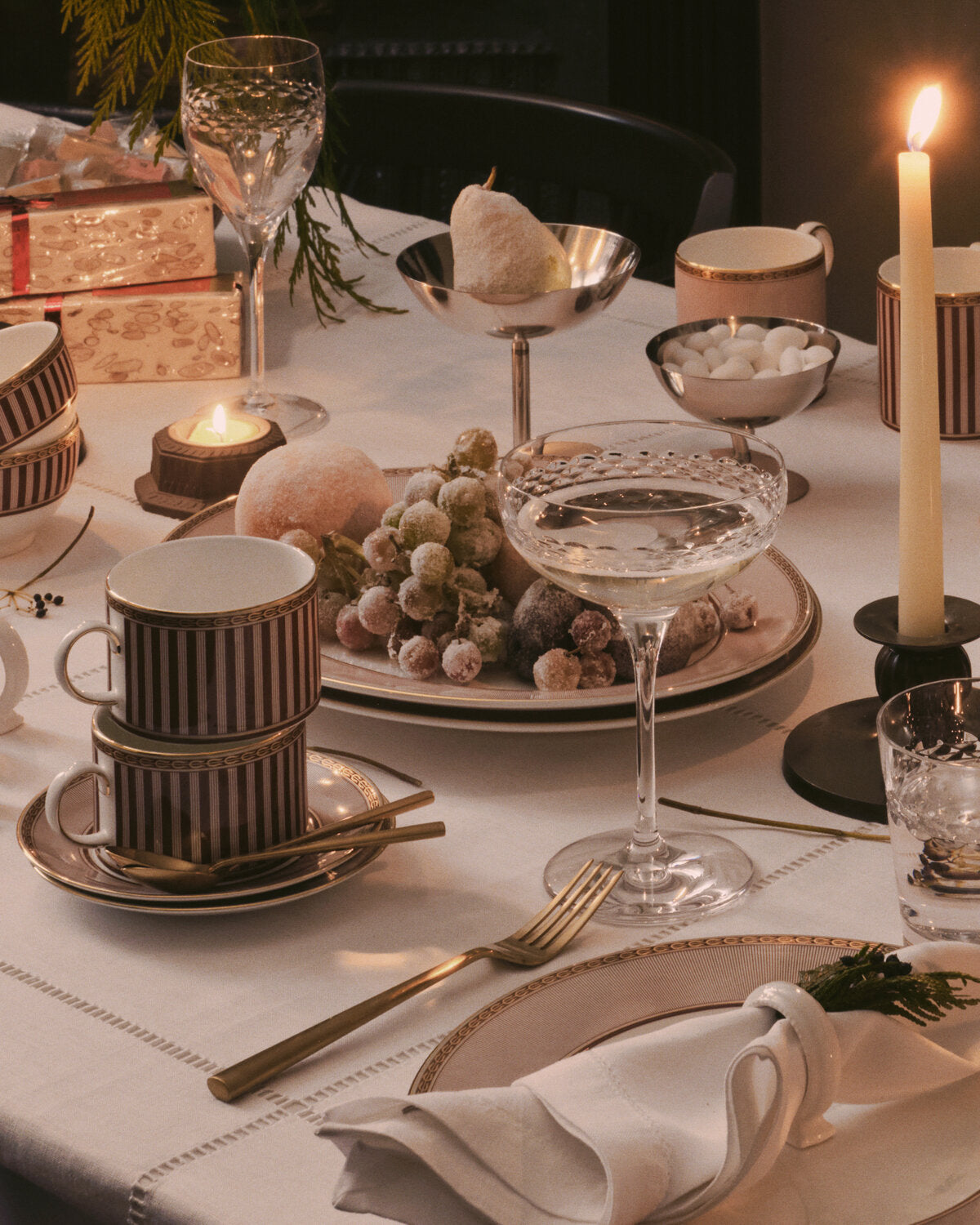 A styled Wedgwood dining table setting with red splendour striped ceramic mugs, gold-toned cutlery, crystal glasses, candles, and a plate of grapes in warm candlelight.