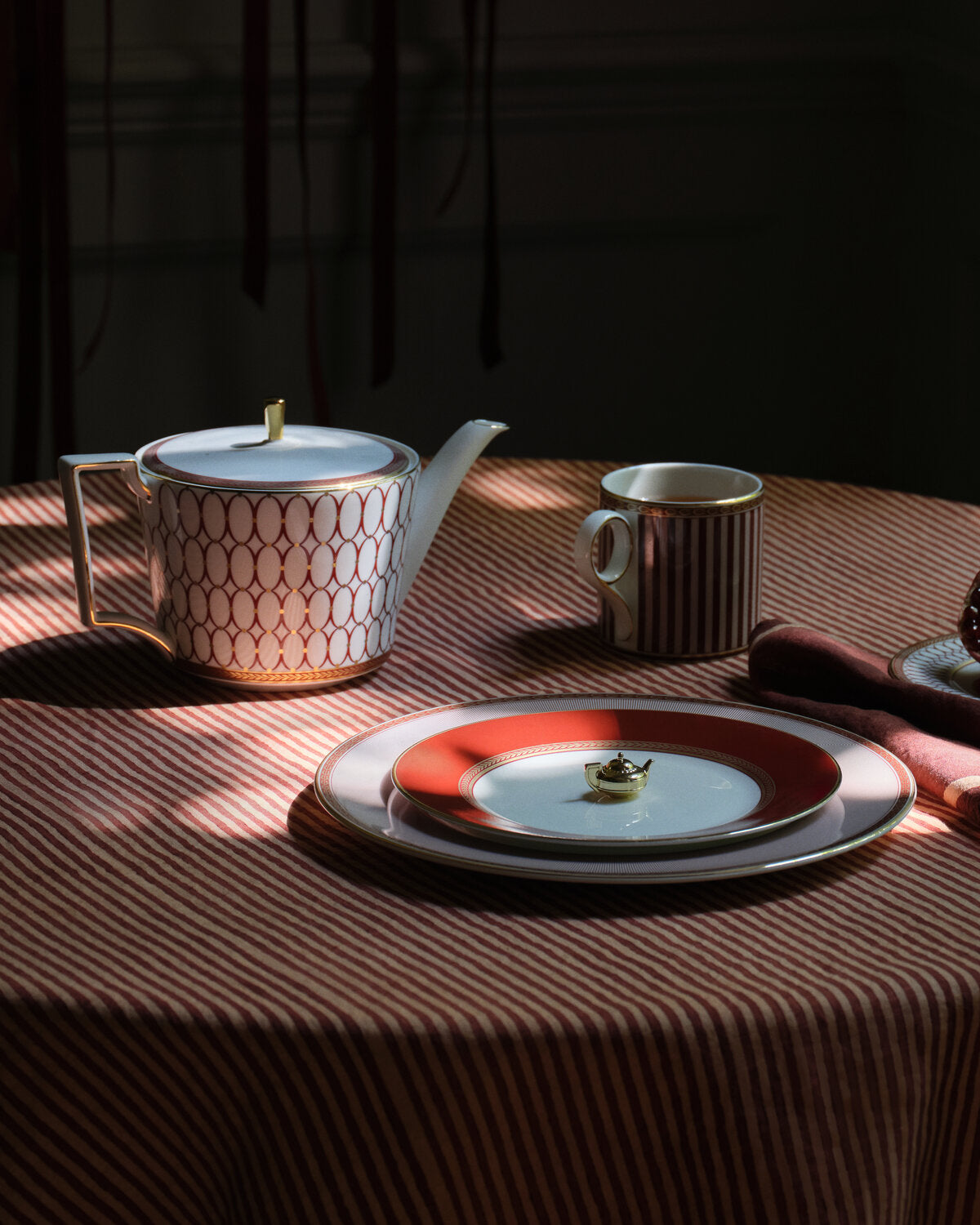 A renaissance red ceramic teapot and matching cup with a striped and geometric pattern, arranged on a striped tablecloth with dramatic sunlight and shadows.