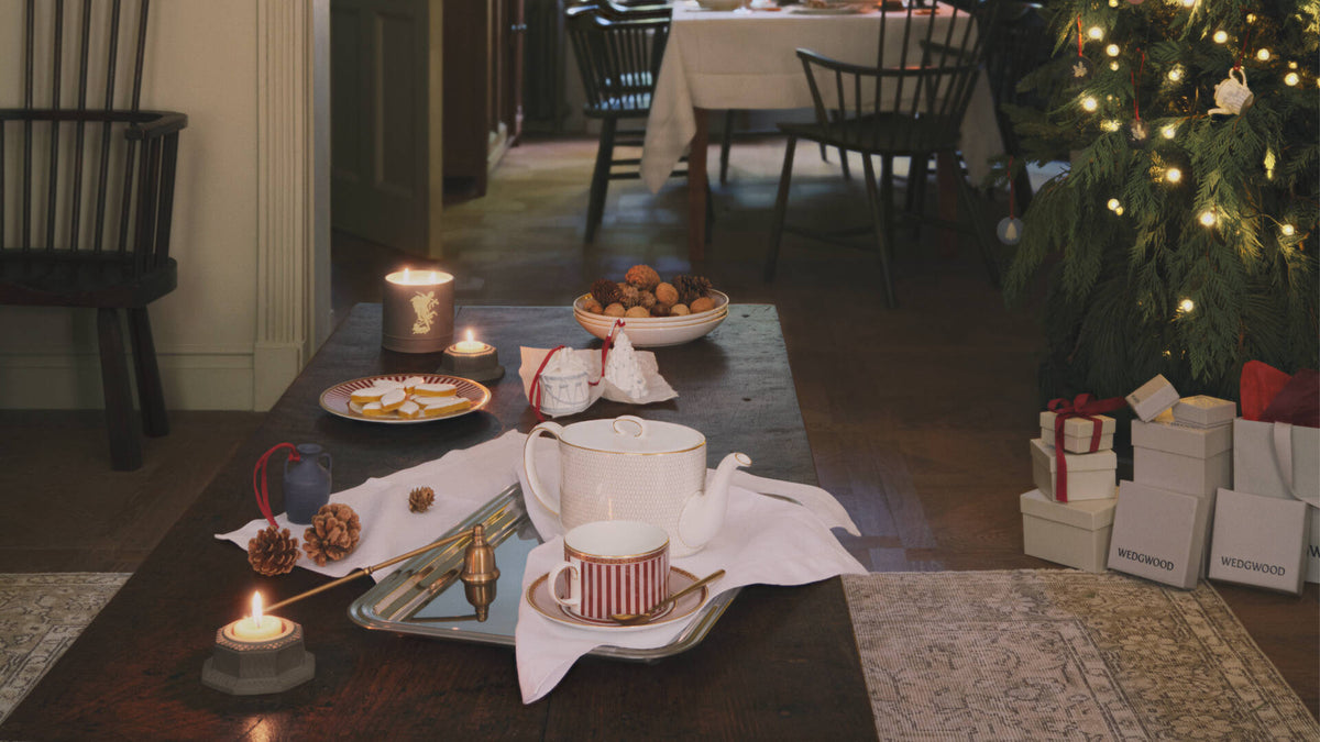 A styled holiday tea setup on a dark wooden table, featuring a Wedgwood white and gold Gio teapot, a white red splendour teacup with red stripes, candles, cookies, and a Christmas tree in the background.