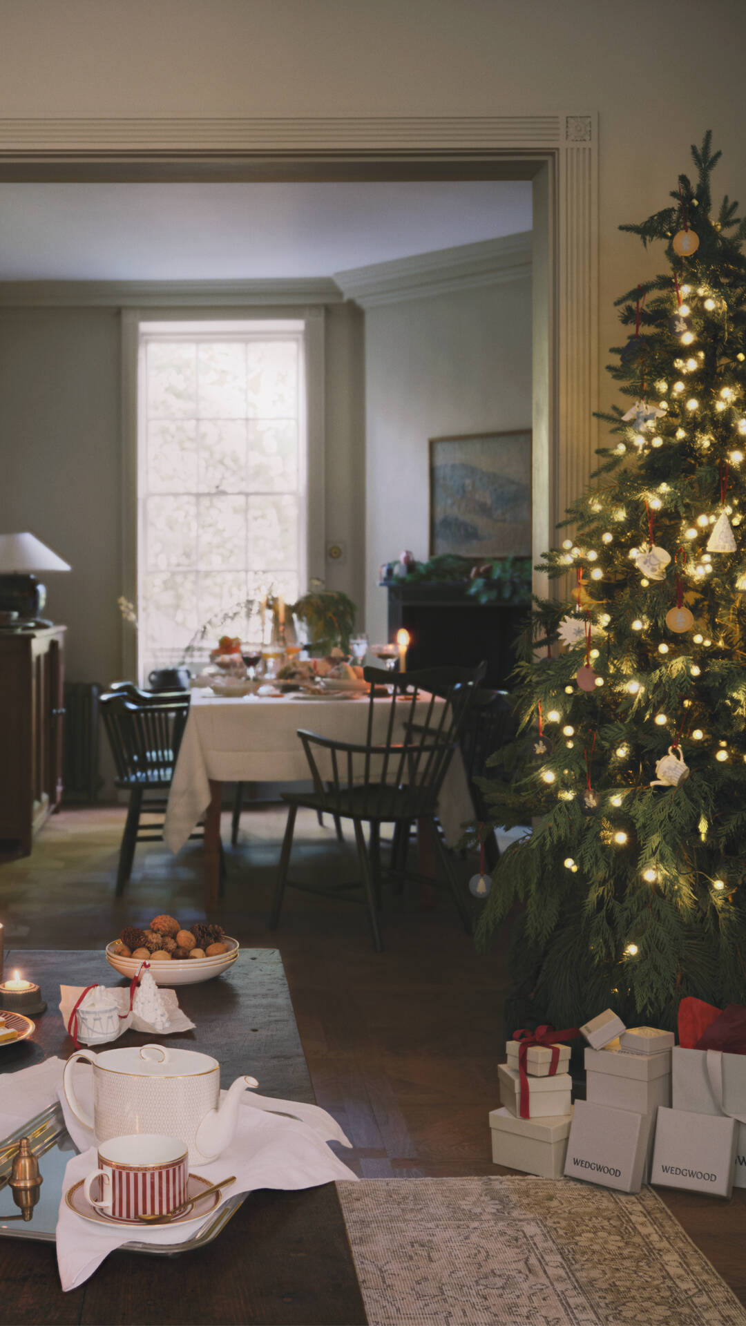 A cozy holiday dining room scene with a decorated Christmas tree on the right, a wedgwood table set with teacups in the foreground, and a dining table in the background. Several white gift boxes with red ribbons and bows are visible near the base of the tree.