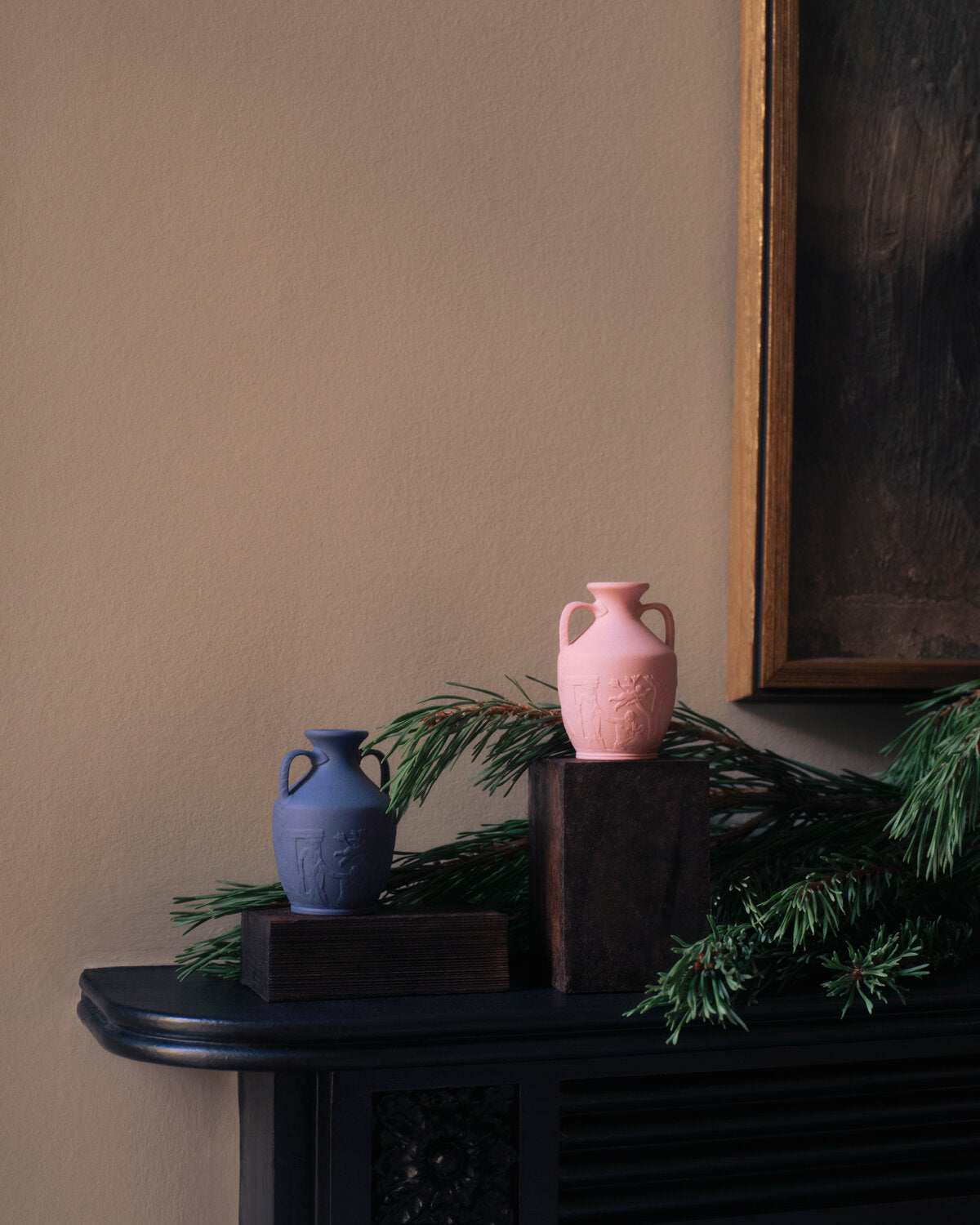 Two decorative mini Portland vases, one dusty pink and one muted blue, displayed on dark wooden blocks atop a dark mantelpiece with pine branches against a warm beige wall.