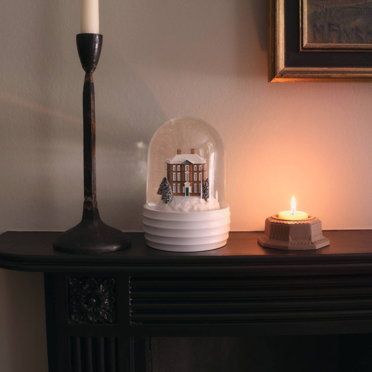 A white ceramic Wedgwood house snow globe under a clear dome sits on a dark mantel beside a lit candle holder and a tall candle, against a warm-toned wall.