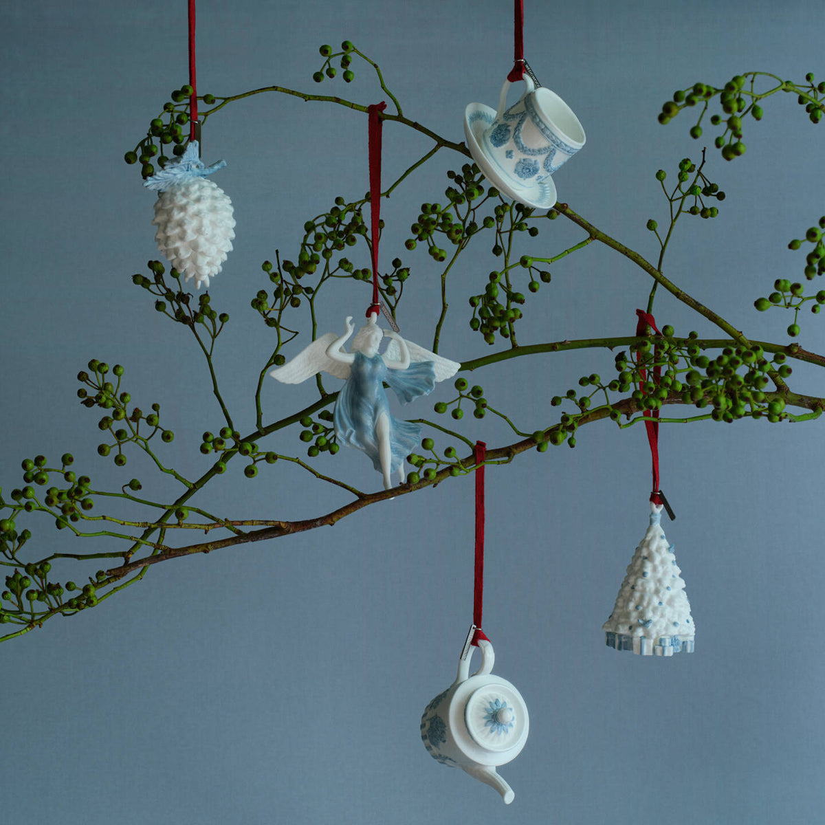 Five ceramic Wedgwood blue and white Christmas ornaments shaped like a pinecone, angel, teapot, teacup, and miniature Christmas tree, hanging from a decorative branch with red strings against a blue-gray background.