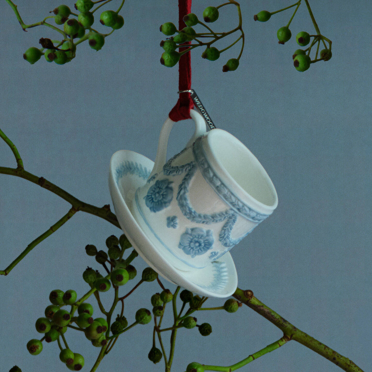 A white and blue ceramic Wedgwood teacup and saucer ornament decorated with a blue floral pattern, hanging from a red ribbon on a tree branch with green berries against a blue-gray background.