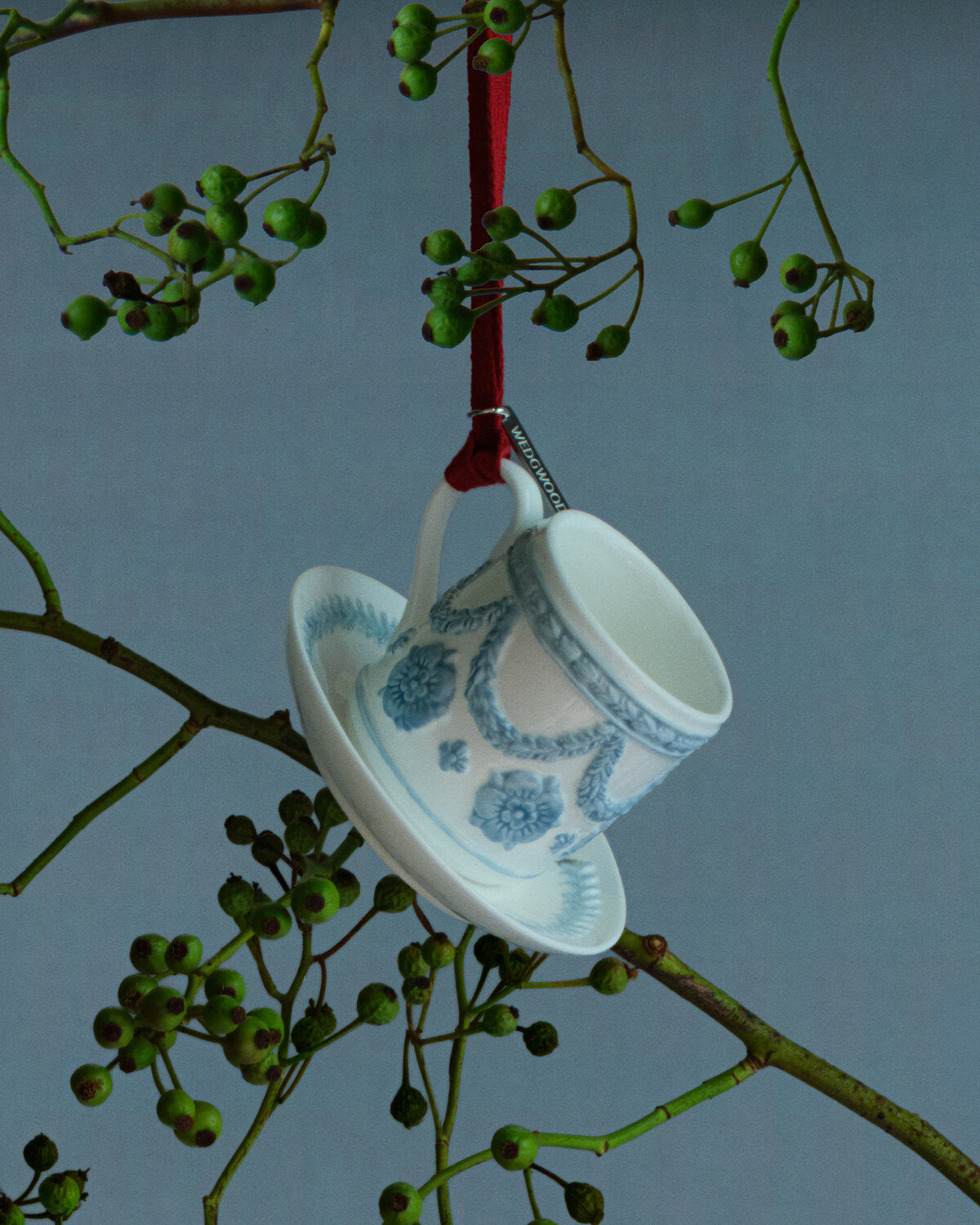 A white ceramic Wedgwood teacup and saucer ornament with blue floral patterns, hanging from a red ribbon threaded through green berries on a tree branch against a gray-blue background.
