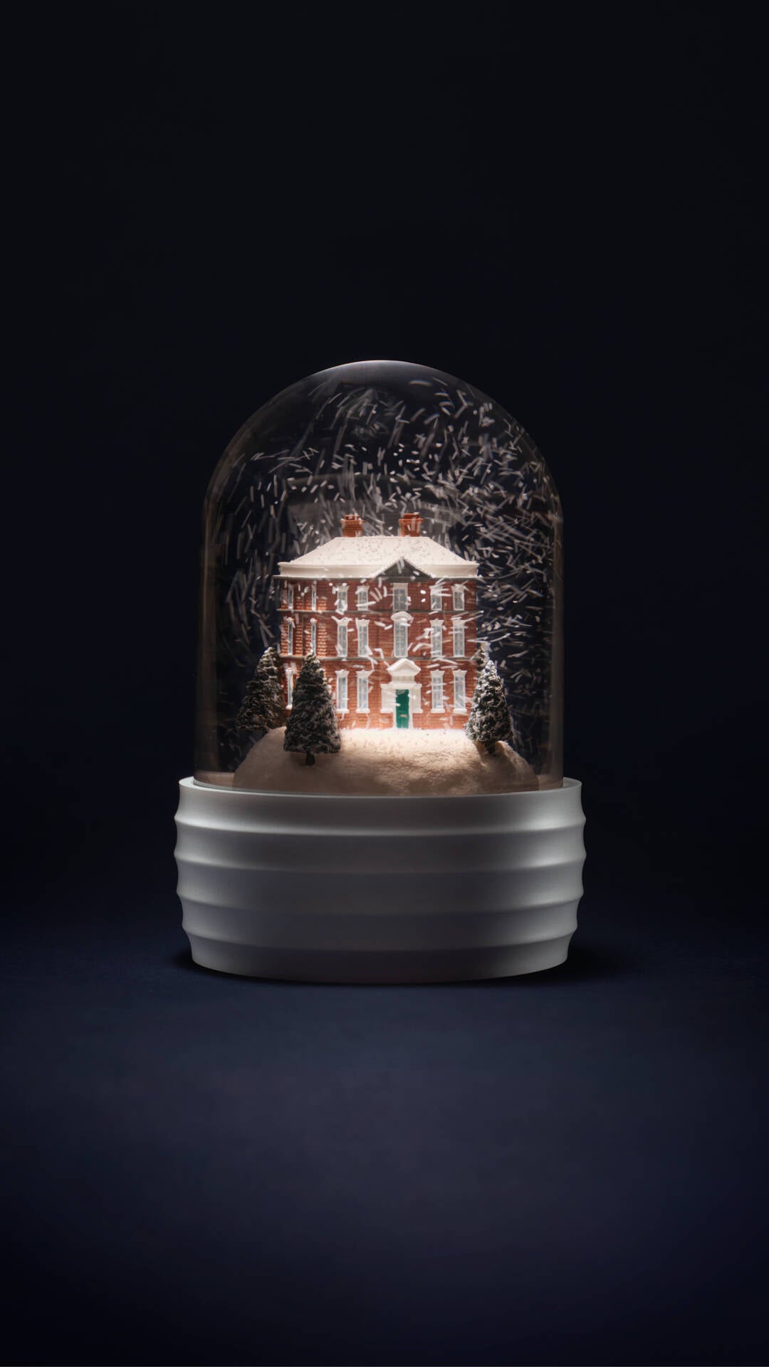 A decorative Wedgwood glass dome snow globe with a miniature lit red brick house, snowy trees, and a white ribbed base, photographed against a very dark navy background.