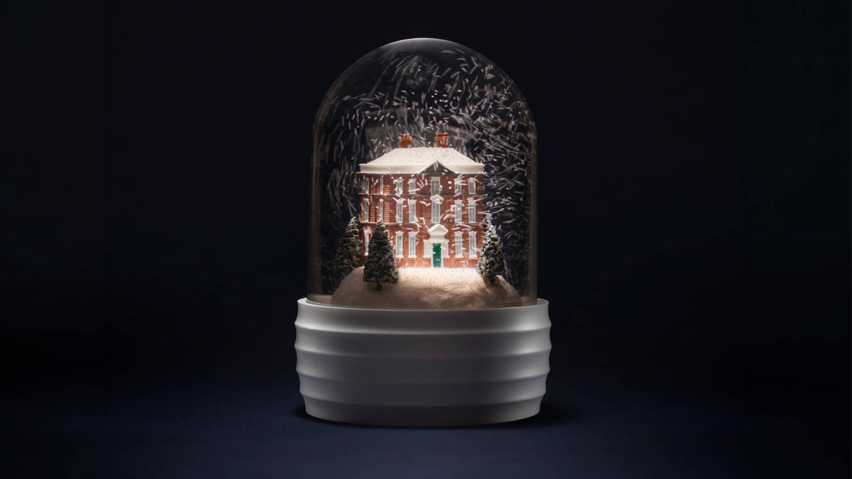 A decorative Wedgwood glass dome snow globe with a white ribbed base, featuring a miniature red brick house, snowy trees, and a small evergreen tree inside, photographed against a dark black background.