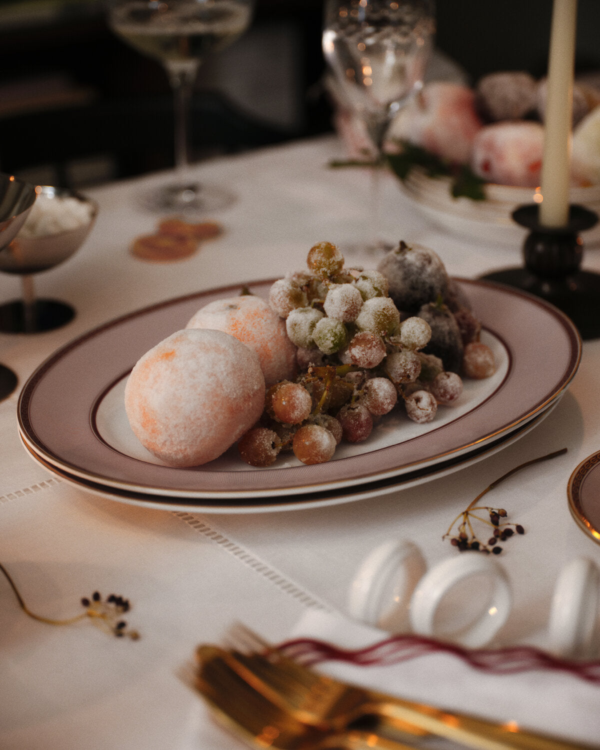 A Wedgwood styled table setting featuring a red splendour plate of frosted grapes and peach-colored spheres on a white tablecloth with gold cutlery and candlesticks in the background.