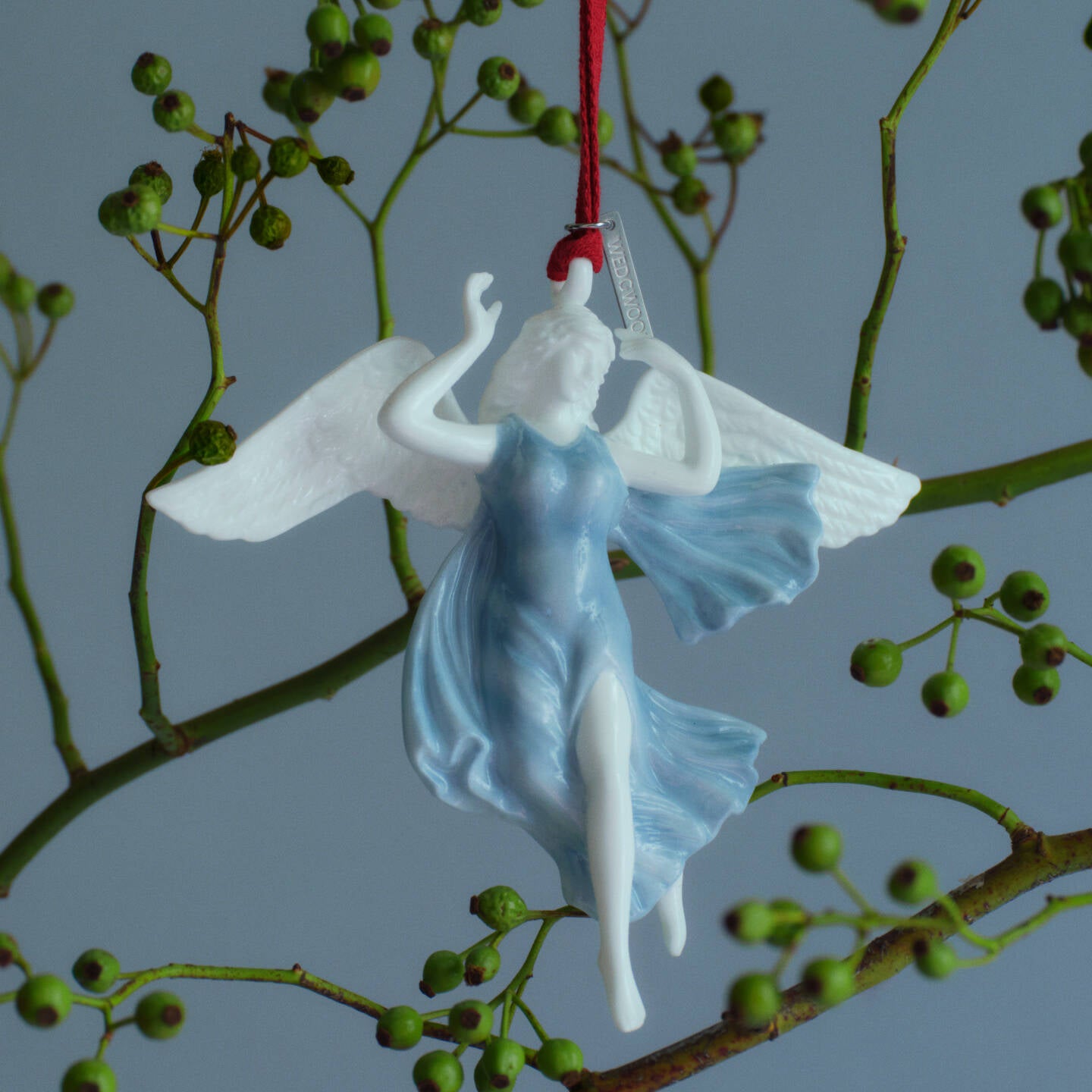 A Wedgwood white and light blue angel Christmas ornament with outstretched wings, hanging from a red string against a gray background with green berry branches.