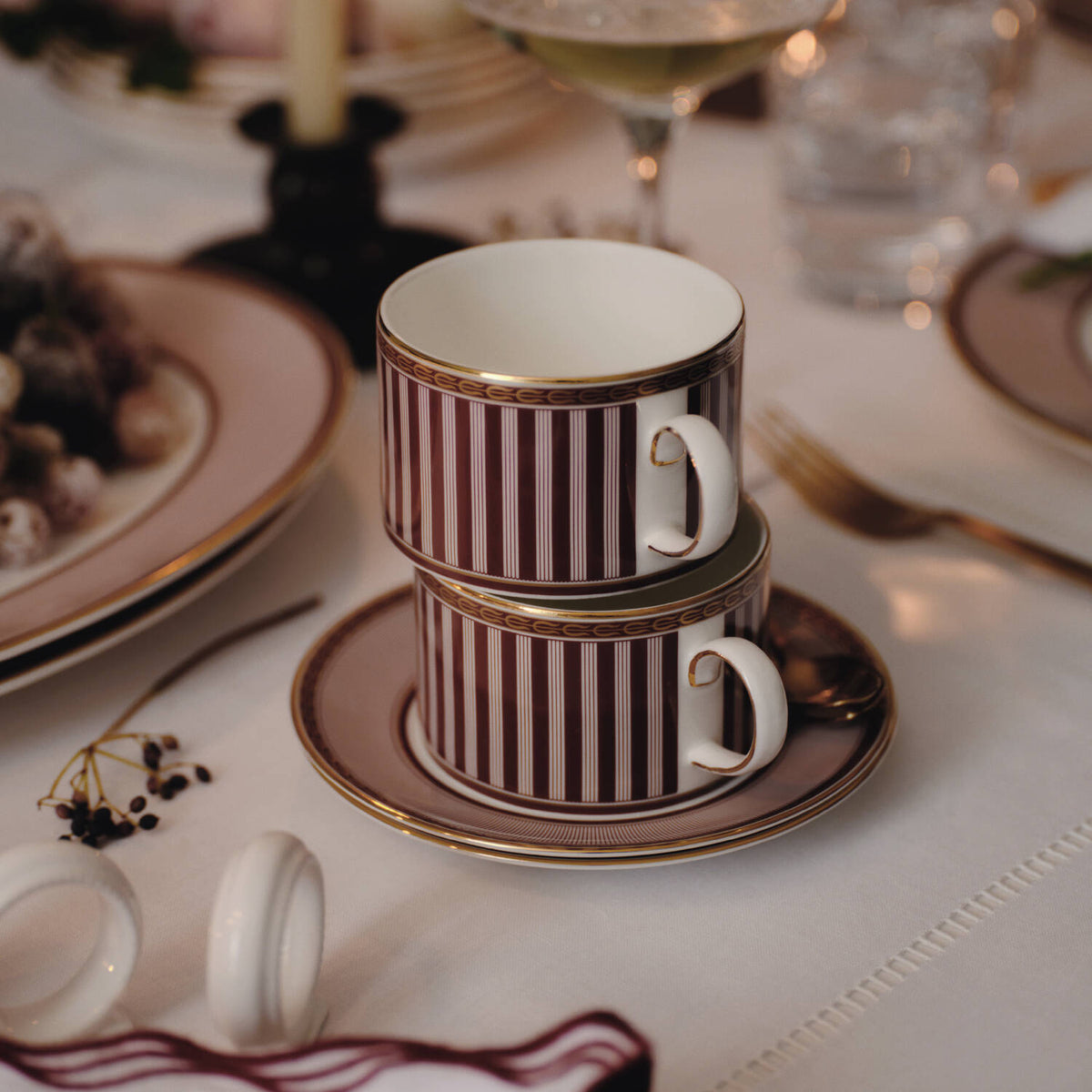 Splendour Red Teacup Saucer Set Of 2 - Two stacked Red Splendour white ceramic teacups with burgundy vertical stripes and gold trim, resting on matching saucers on a white tablecloth at a dinner table.