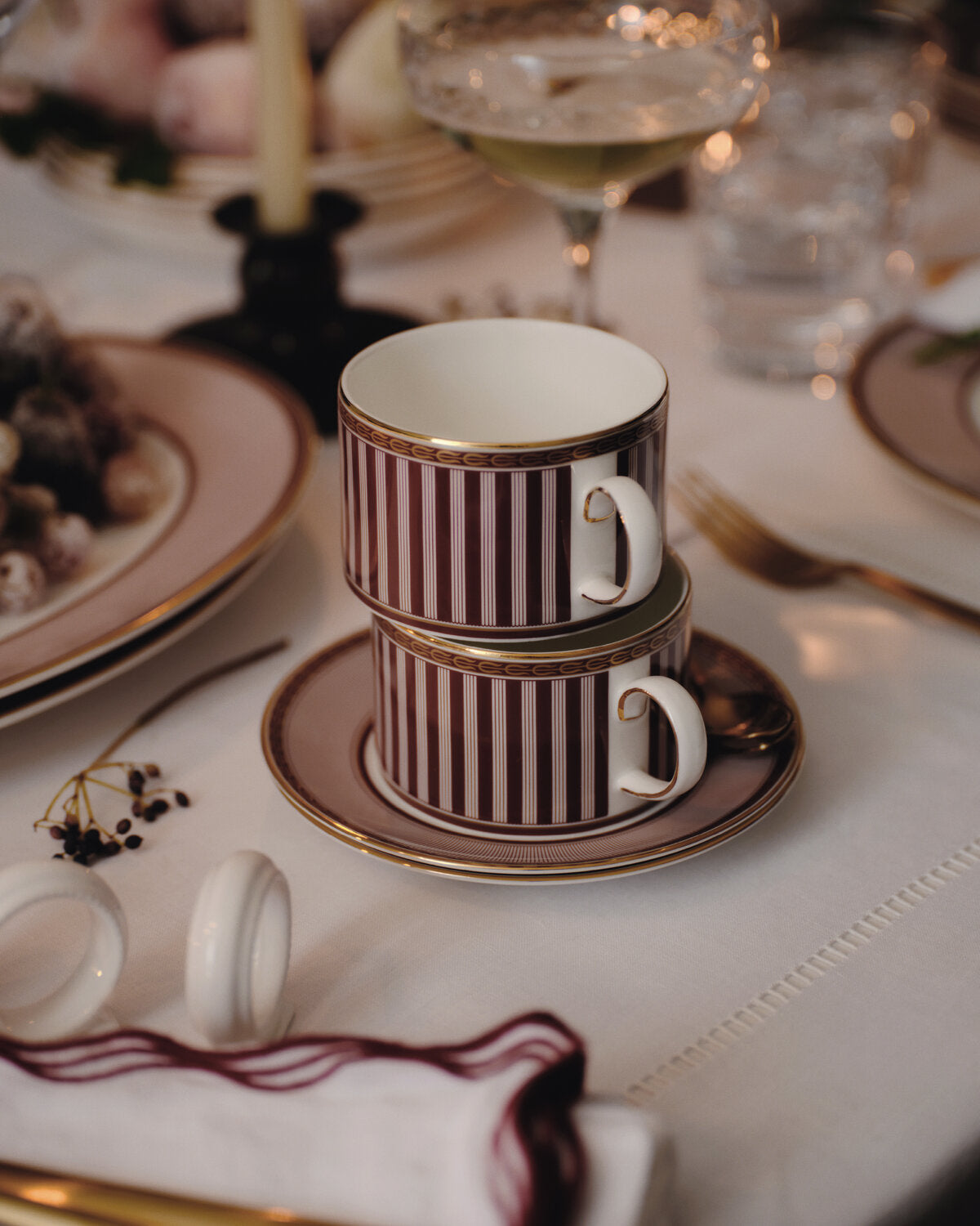 Two stacked wedgwood white porcelain teacups with dark burgundy stripes and gold trim, resting on matching saucers on a white tablecloth at a dinner table.
