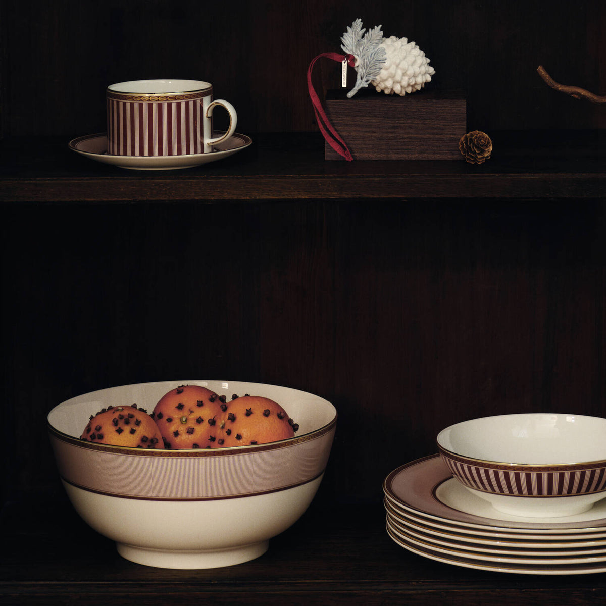 Splendour Red Serving Bowl - A set of elegant Red Splendour ceramic tableware displayed on a dark wooden shelf, including a striped cup and saucer, a large bowl with a beige band, a stack of plates, and a matching small bowl.