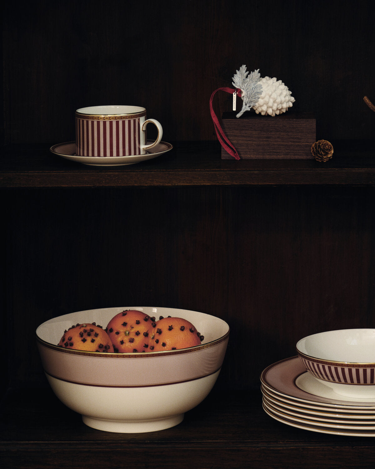 A collection of elegant red splendour ceramic tableware displayed on dark wooden shelves, featuring a striped cup and saucer, a large bowl with a solid beige band, a stack of matching plates, and a small bowl on a saucer, styled with decorative fruit and a pinecone ornament.