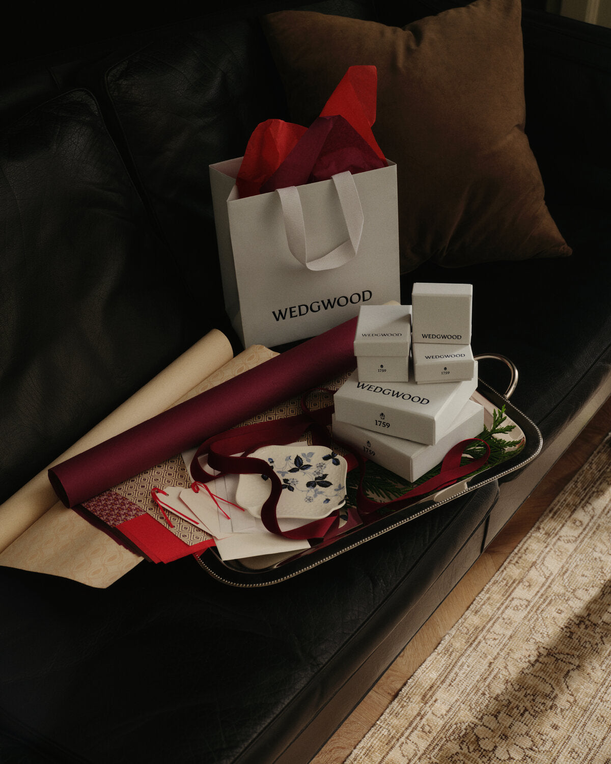 Wedowood gift set arranged on a black leather sofa, including a white branded shopping bag with red tissue paper, several white product boxes, a rolled burgundy fabric item, and folded gift papers on a tray.