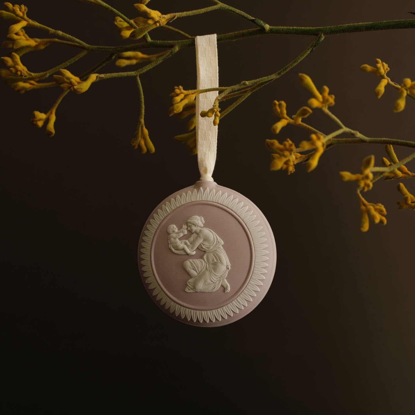 A round pale pink and white Wedgwood jasperware decorative hanging ornament with a relief figure design, suspended from a cream ribbon against a dark background with yellow flowers above.