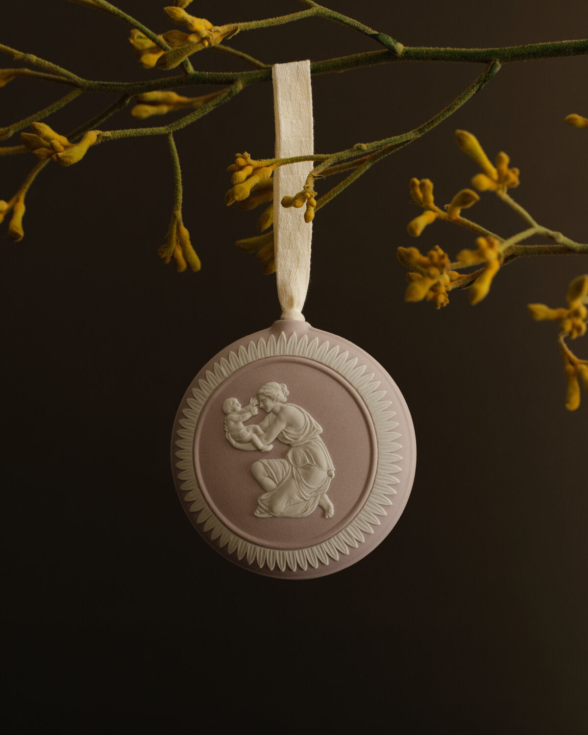 A Jasperware round pale mink and white decorative hanging ornament with an embossed classical figure motif, suspended from a white ribbon against a dark background with yellow flowers above.