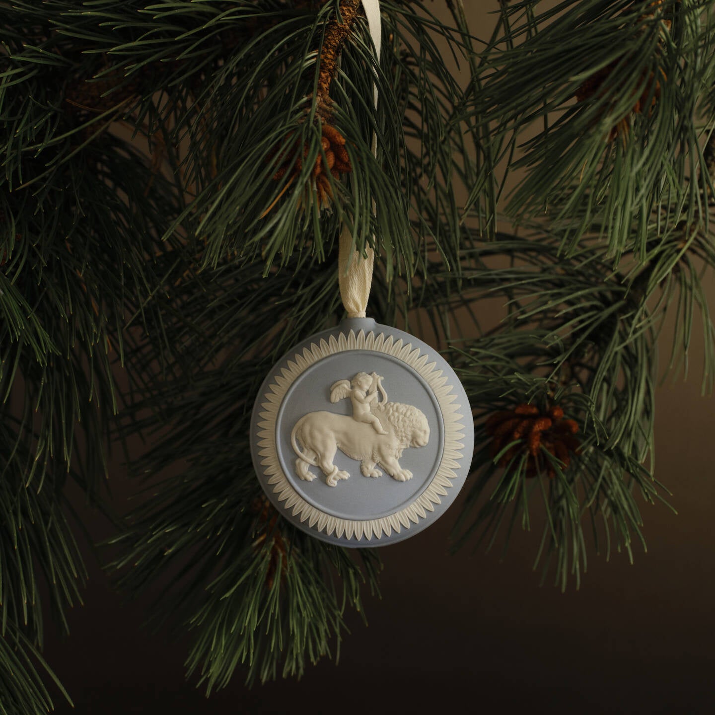 A round decorative hanging Wedgwood jasperware ornament with a lion and lamb motif, suspended from a green pine tree branch against a dark brown background.