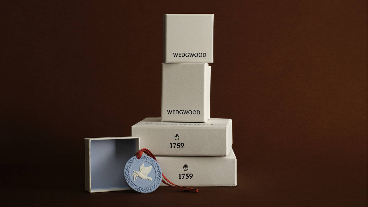 Wedgwood 1759 box set featuring three stacked white wooden boxes of varying heights, with a matching open box and a Jasperware round blue medallion tag with a white dove and red ribbon, set against a solid dark brown background.