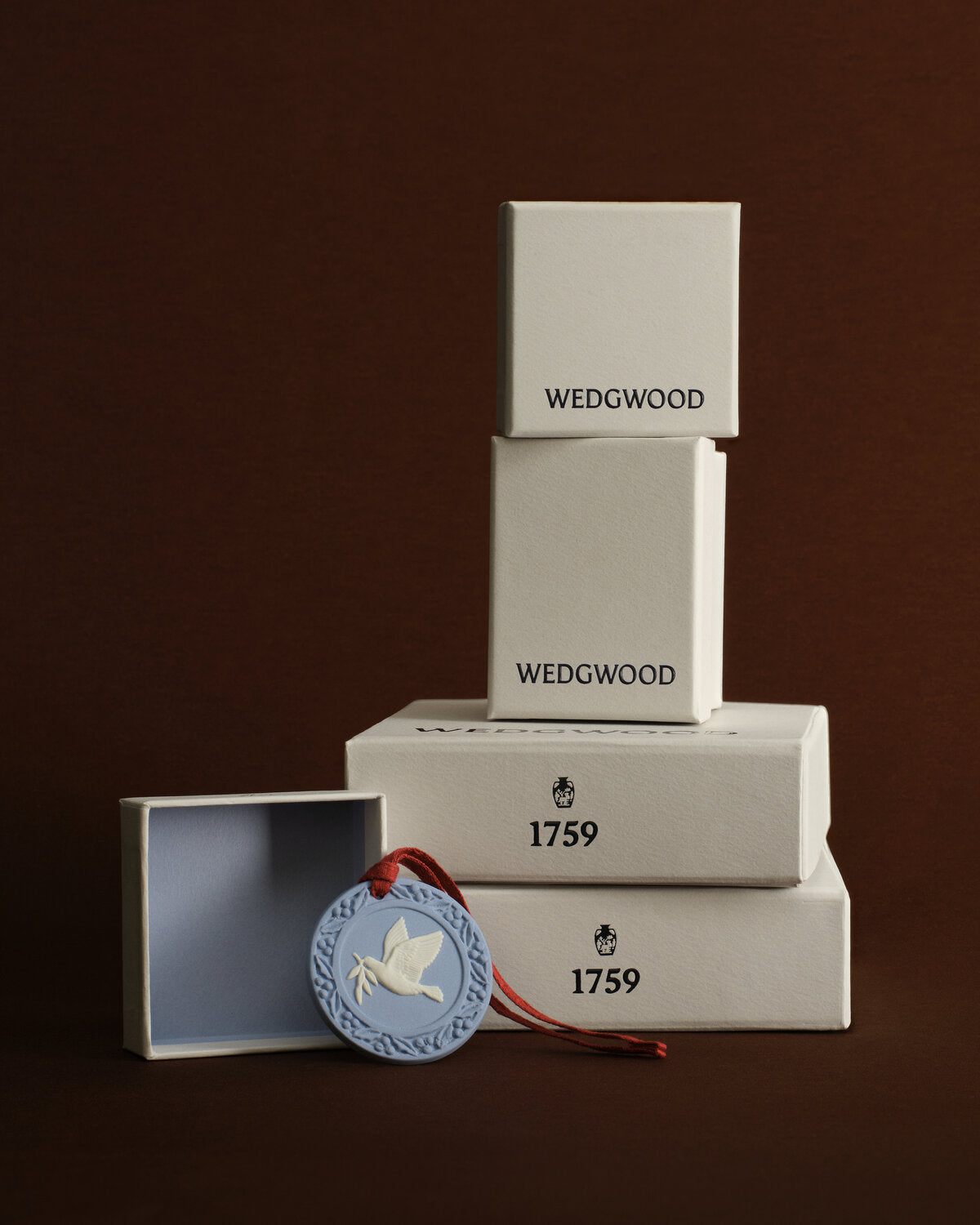 A stacked set of Wedgwood presentation boxes in off-white wood, with one open box and a round blue decorative tag featuring a blue and white  jasperware ornament with dove design and red ribbon, set against a dark brown background.
