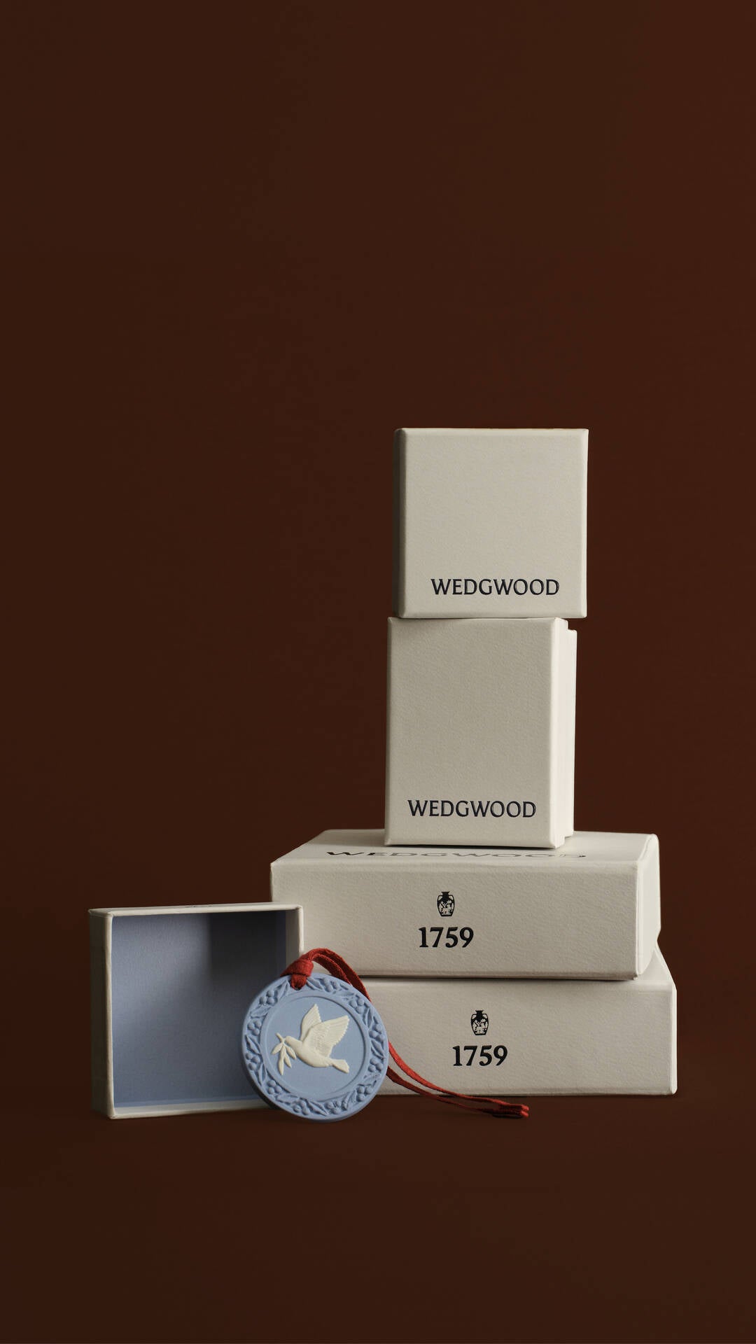 Wedgwood 1759 box set featuring three stacked white wooden boxes of varying heights, with a matching open box and a Jasperware round blue medallion tag with a white dove and red ribbon, set against a solid dark brown background.
