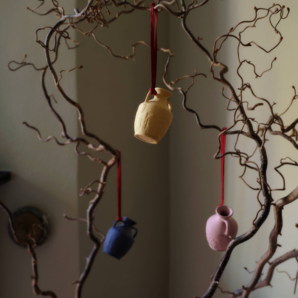 Three small hanging Wedgwood jasperware Portland vase cream, blue, and pink, suspended from red ribbons among bare brown branches against a muted indoor background.