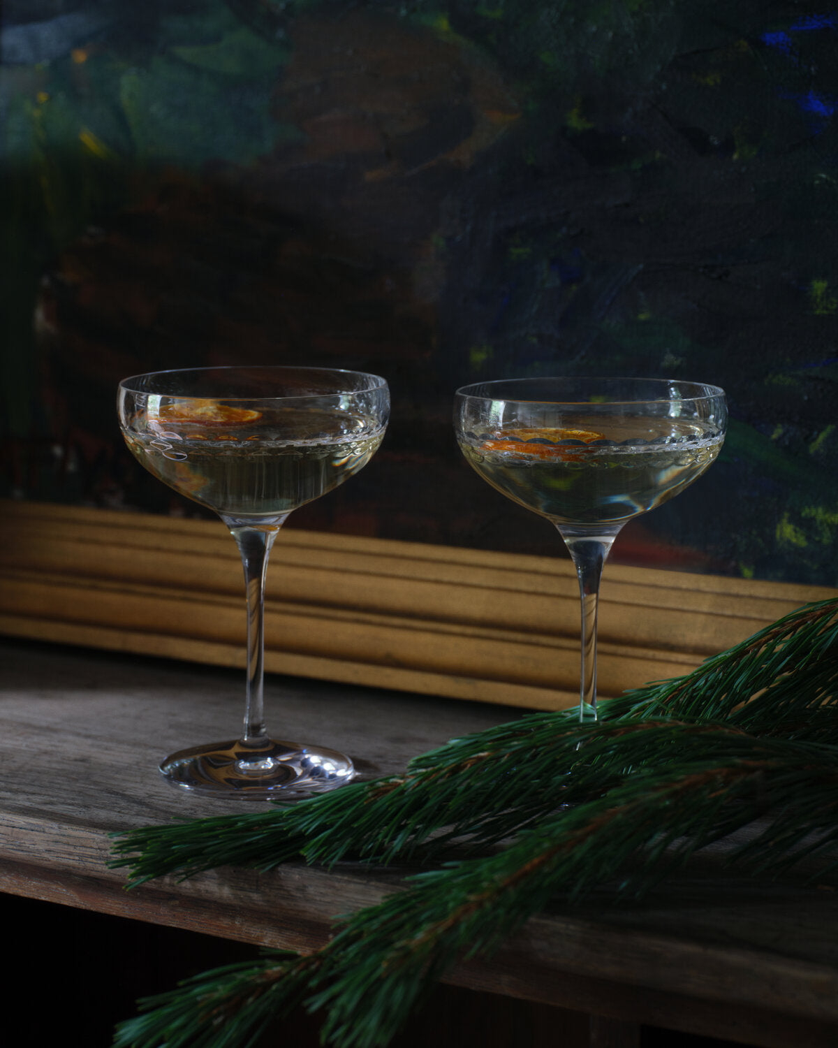 Two coupe glasses for the wedgwood Neo collection filled with a pale golden drink and orange slices, set on a rustic wooden surface with pine branches in the foreground and a dark painted background.