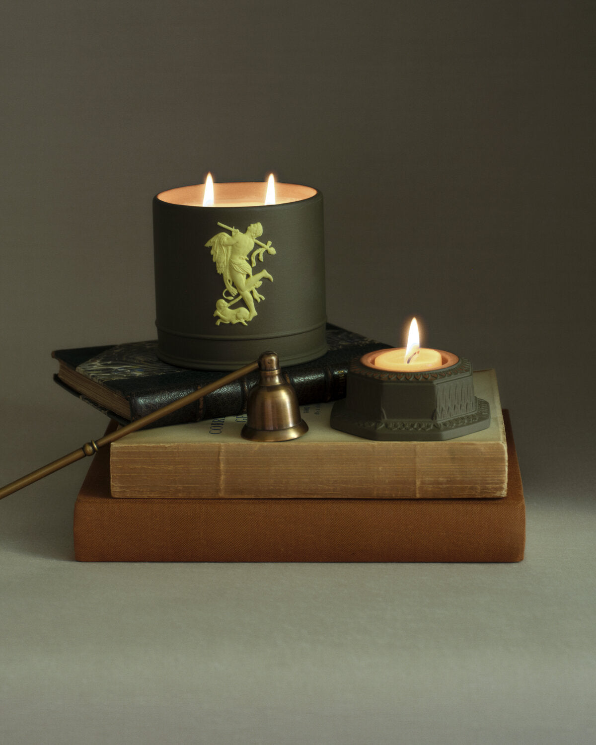 A styled product shot featuring a large Wedgwood jasper Bacchus black candle with a green classical figure relief on the front, lit and resting on a stack of vintage books alongside a small tealight holder and a brass bell.