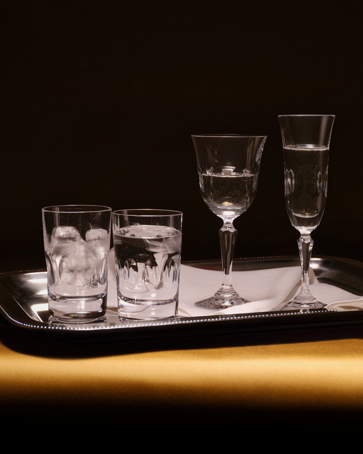 A set of four clear glassware from the Wedgwood Bacchus Crystal collection arranged on a silver tray against a dark background. The set includes two short tumblers with ice, one stemmed goblet, and one tall fluted champagne flute, all with water or clear liquid.
