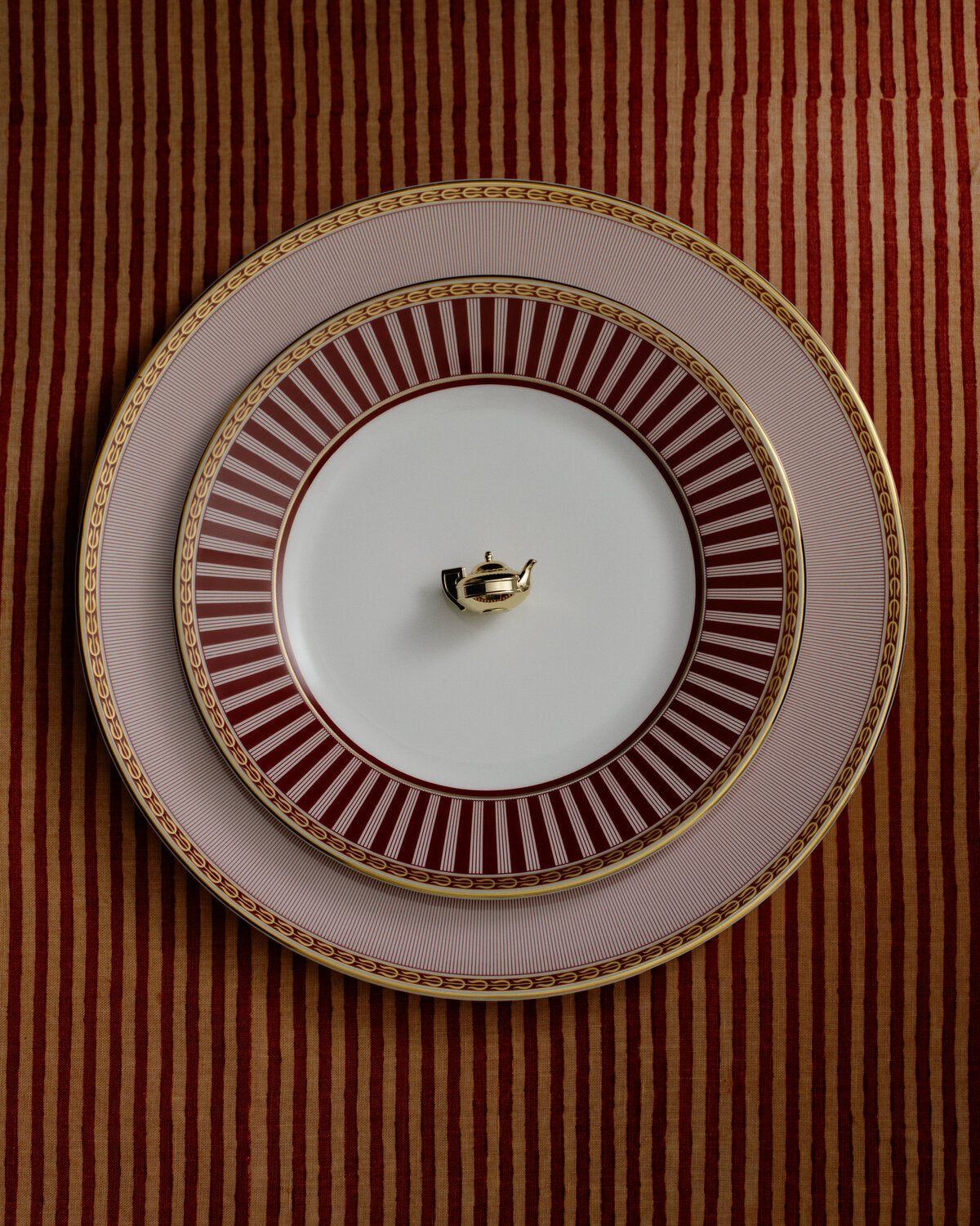 Three stacked ceramic plates from the wedgwood splendour red collection with a decorative gold and red striped pattern, featuring a small gold teapot-shaped ring holder in the center, set against a red and gold striped fabric background.