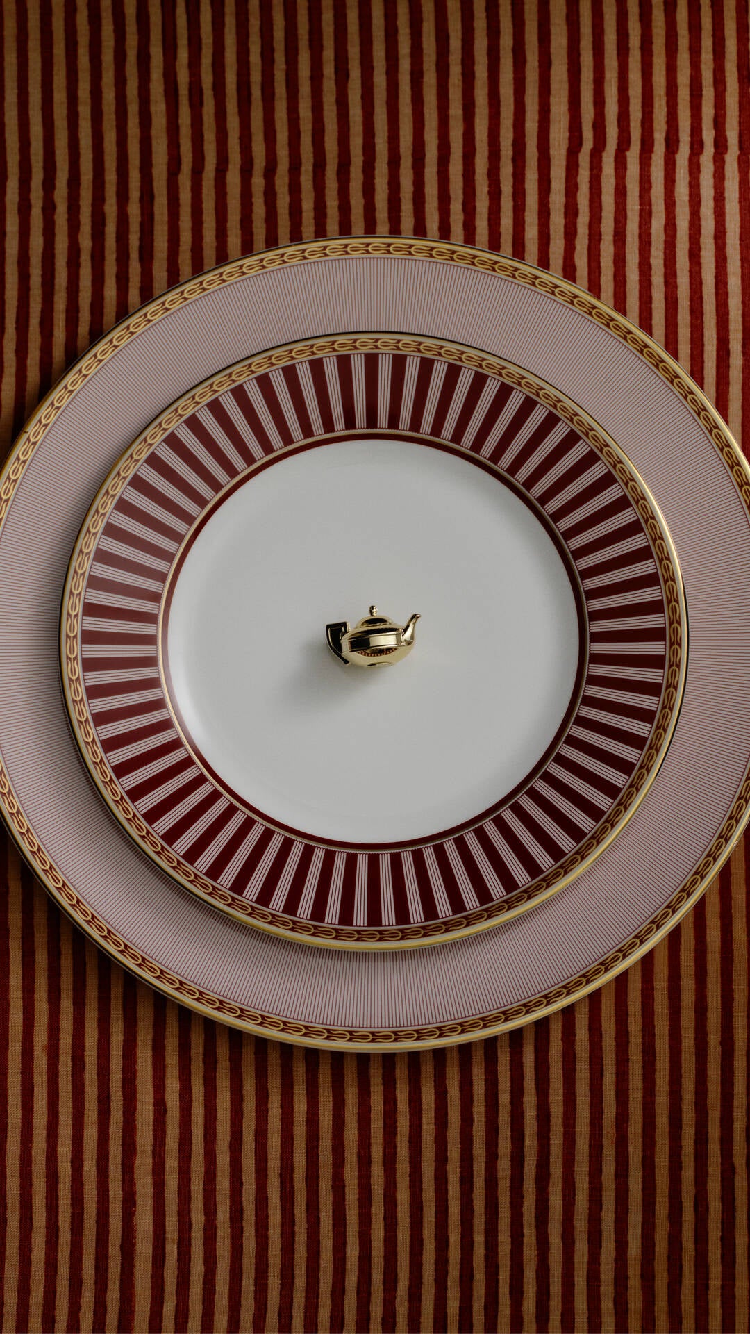 Stacked Red splendour ceramic plates with a white center, a dark red and white sunburst pattern, and a gold rim, set against a red and gold striped background. A small gold teapot is placed in the center of the top plate.