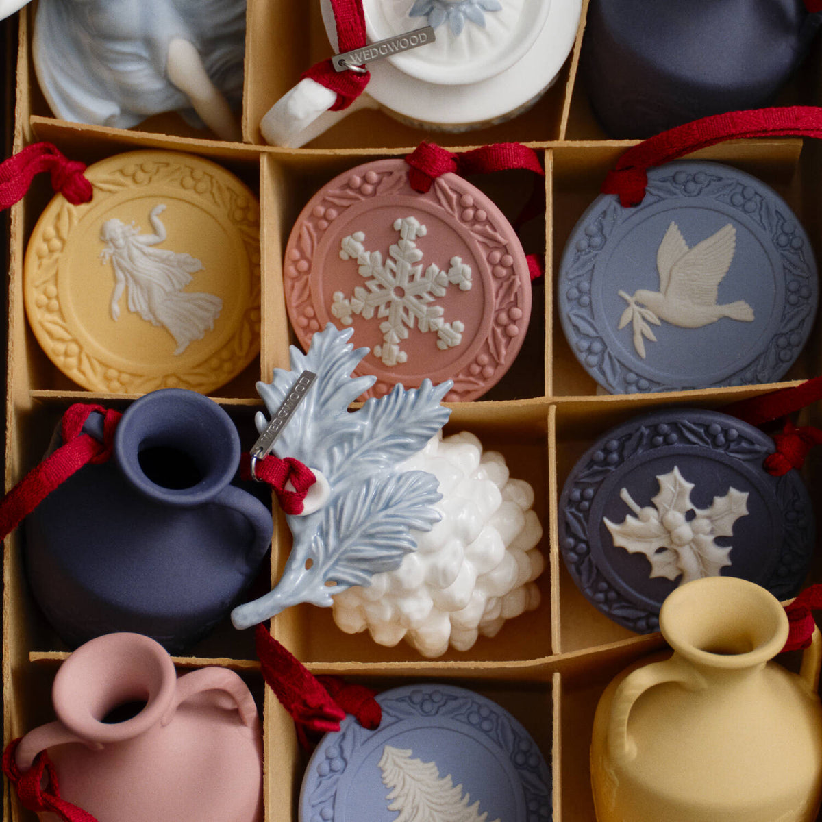 A boxed assortment of small decorative Wedgwood jasperware ornaments and vases in muted pastel colors, each with a red ribbon loop and embossed holiday motifs. The set includes round medallions, a blue pitcher, a pink vase, a pale blue leaf ornament, a white grape cluster ornament, and other matching pieces arranged in a cardboard grid.
