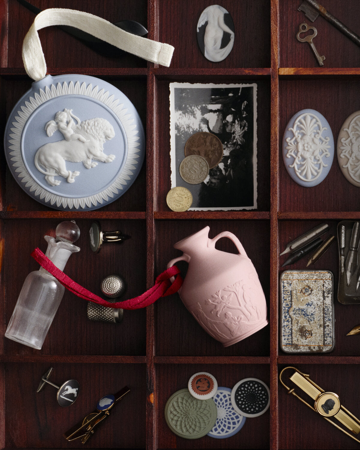 A styled flat-lay of assorted vintage and antique items arranged in a dark wooden compartment organizer, including a wedgwood jasper pale blue and white ceramic medallion with a lion relief, a pink embossed portland vase with a red ribbon, keys, coins, buttons, tools, and small decorative objects.