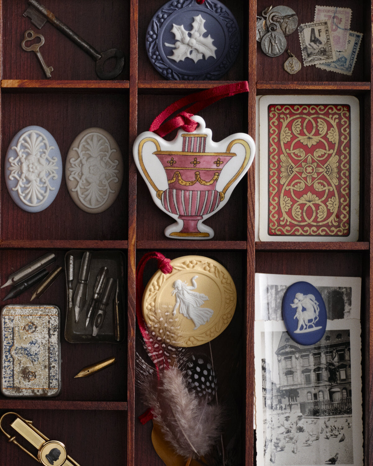 A styled flat-lay of vintage-inspired decorative items and Wedgwood Christmas decorations arranged in a dark wooden grid, including a pink and gold Portland vase ornament with a red ribbon, a round gold medallion, blue and white brooches, old keys, stamps, feathers, and antique-style cards.