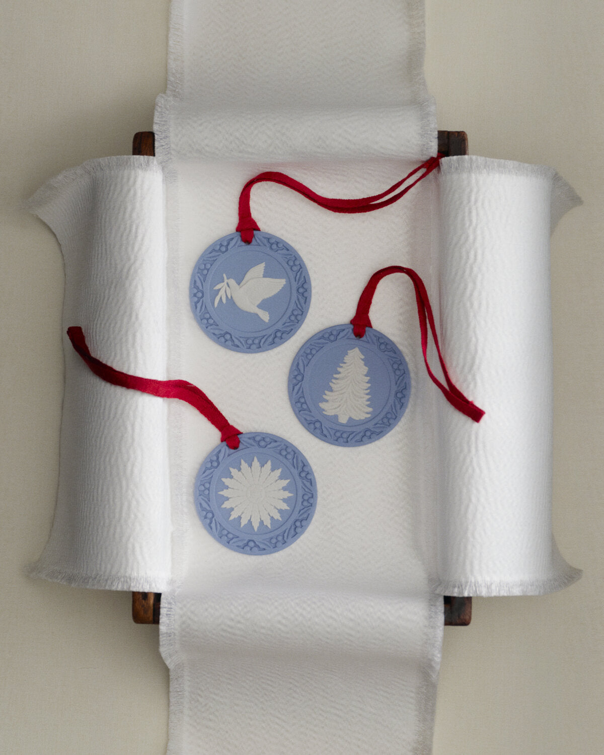 Three round Wedgwood blue jasperware Christmas ornaments with red hanging ribbons are displayed on a white ceramic ribbon runner. The ornaments feature white cutout designs: a dove, a Christmas tree, and a star of David.