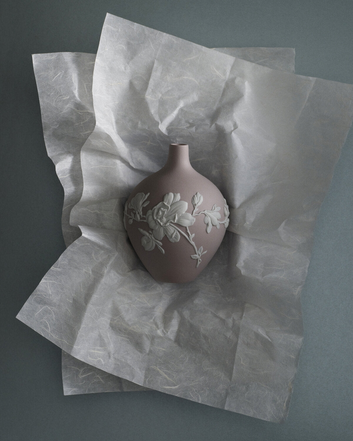 A small Wedgwood matte taupe jasperware vase with white embossed floral magnolia detailing, resting on crumpled translucent tissue paper against a dark gray background.