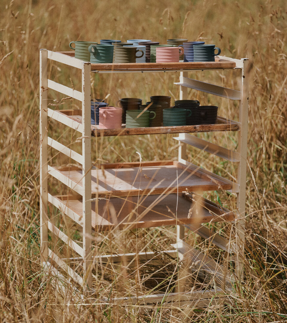 A rustic white metal and wood shelf rack filled with colorful JW Anderson jasperware mugs and bowls, set outdoors in a field of dry golden grass.