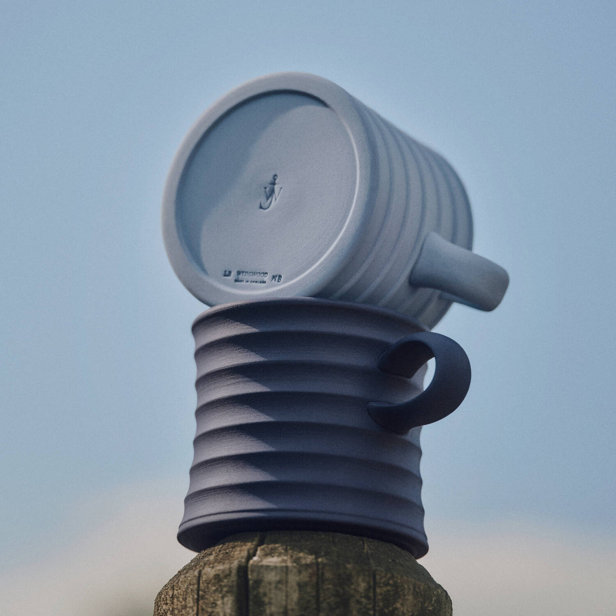 Two ribbed JW Anderson jasperware mugs stacked on a wooden post against a pale blue sky. The top mug is light blue-gray and shown upside down, while the bottom mug is a darker navy blue and standing upright.