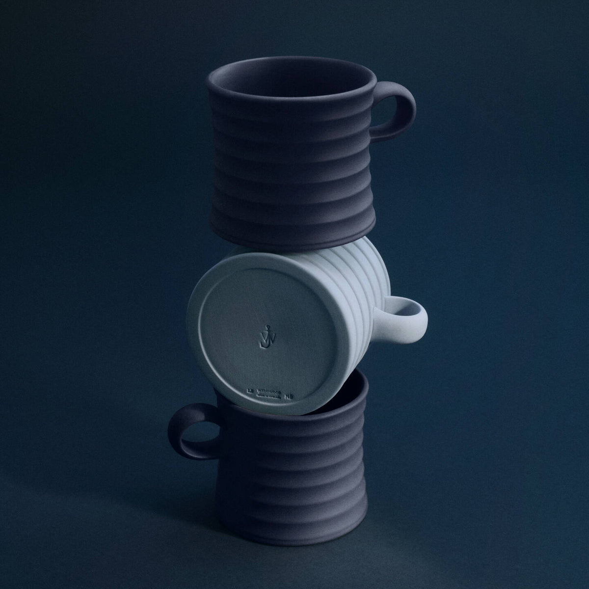 JW Anderson Mug Pair Mercury / Jasper Blue - Three ribbed JW Anderson jasperware mugs stacked in a tower on a dark navy background. The top and bottom mugs are dark charcoal gray, and the middle mug is a light gray.