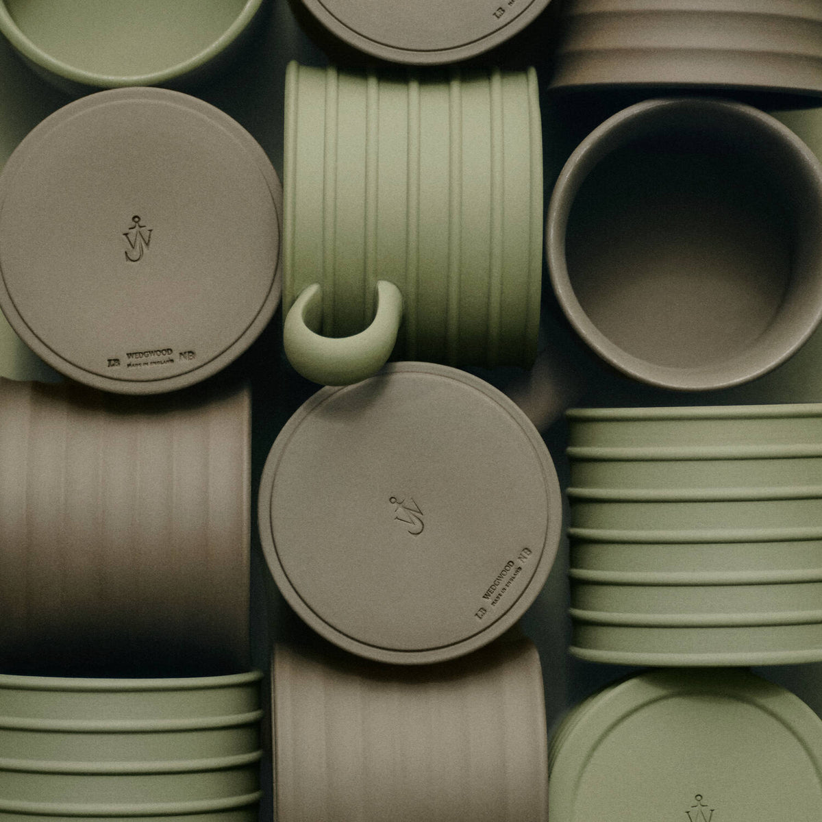 A close-up, top-down view of a collection of matte JW Anderson jasperware mugs and matching lids in a muted sage green and warm taupe-beige colorway. The mugs feature a unique C-shaped handle on one piece, and the lids are embossed with a small logo and the brand name 'monmouth'.