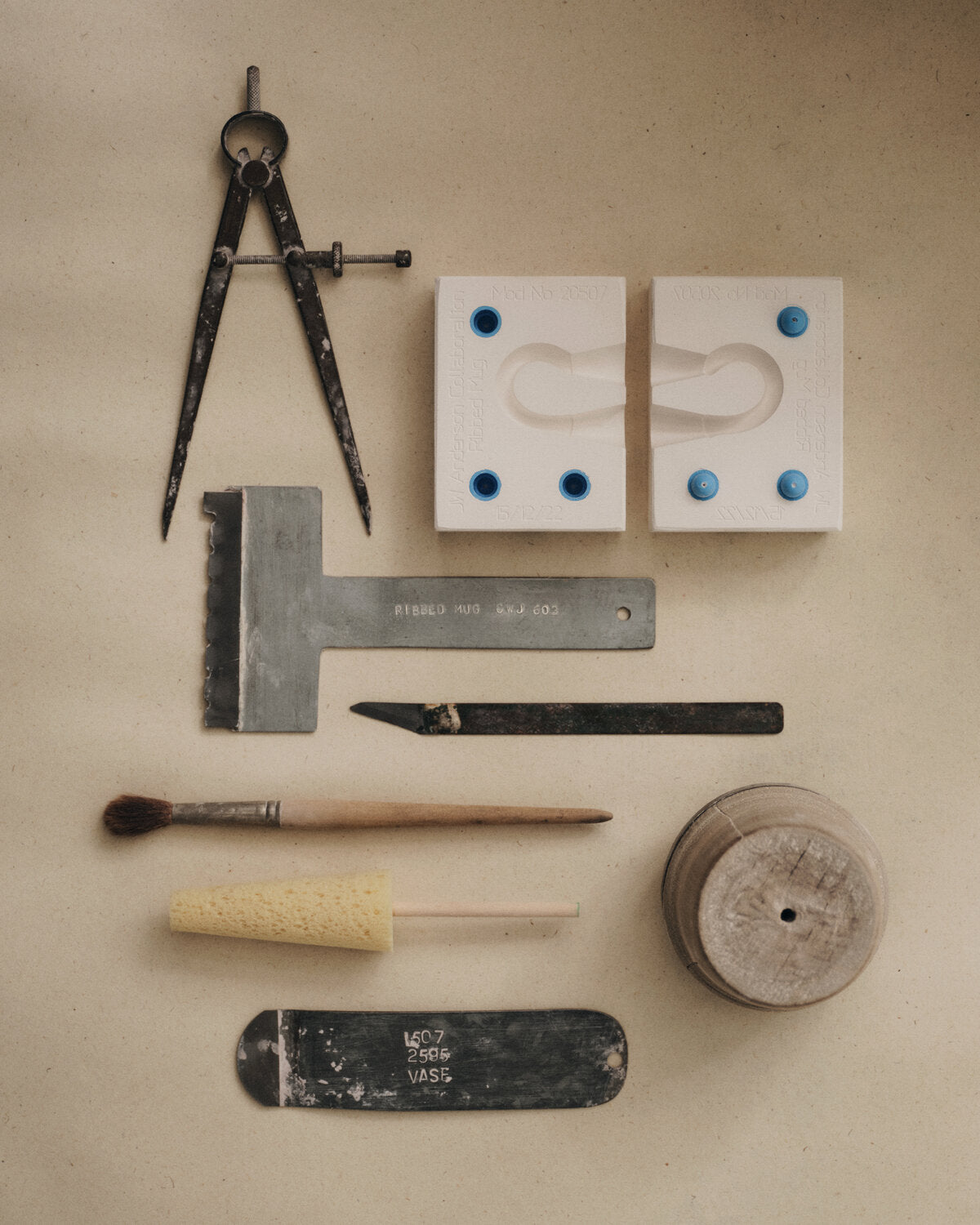 A flat-lay arrangement of various ceramic and pottery tools on a textured beige background, including a metal compass, two white ceramic molds, a metal square, a sculpting knife, a paintbrush, a sponge, and a wooden roller.