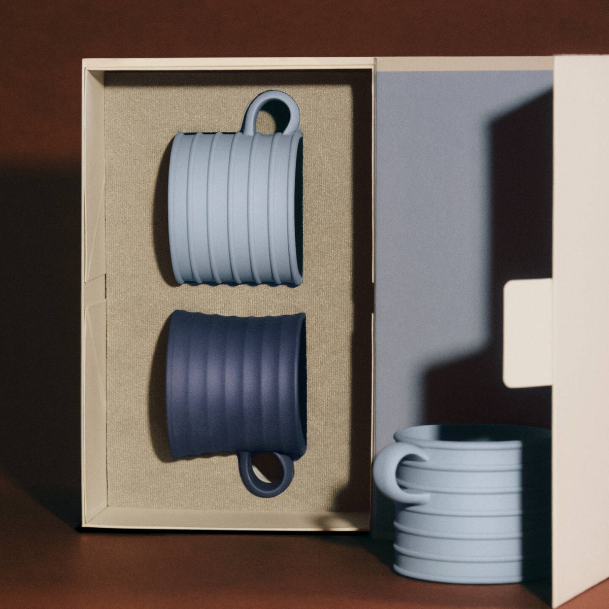 JW Anderson Mug Pair Mercury / Jasper Blue - Two ribbed JW Anderson jasperware mugs with loop handles, one in pale blue and one in dark navy, presented inside a beige gift box with additional mugs stacked outside on a dark brown surface.
