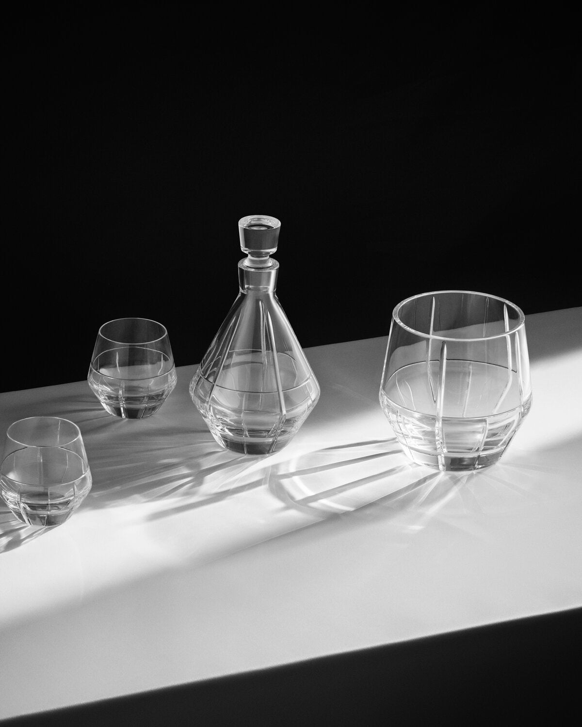 A Vera Wang clear glass decanter and two matching lowball glasses arranged on a white surface against a black background, lit with dramatic directional sunlight that creates strong shadows.